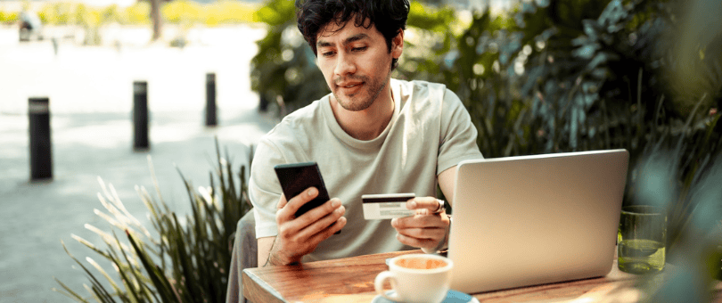 man shopping on his laptop at a cafe, checking his phone perhaps for an OTP to illustrate Signifyd 3D Secure explainer